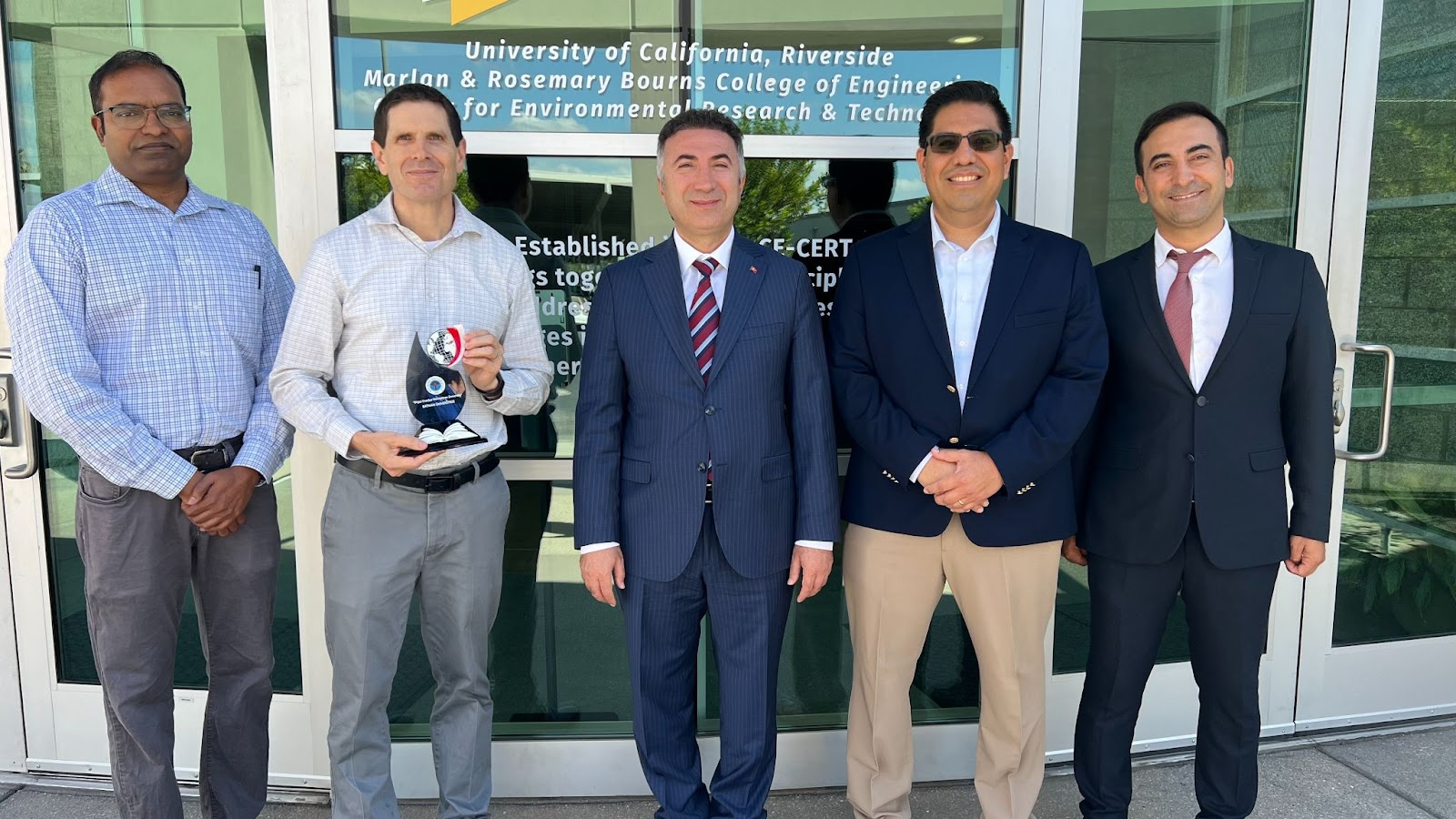 A photo of five men standing in a line outside of the CE-CERT building at UC Riverside. From left to right: Arun Raju, Don Collins (holding a plaque), Prof. İdris Demir, Alfredo Martinez-Morales, and Musa Yilmaz. The photo was taken during Prof. Demir's visit to explore opportunities for international collaboration in energy research and academic exchange.