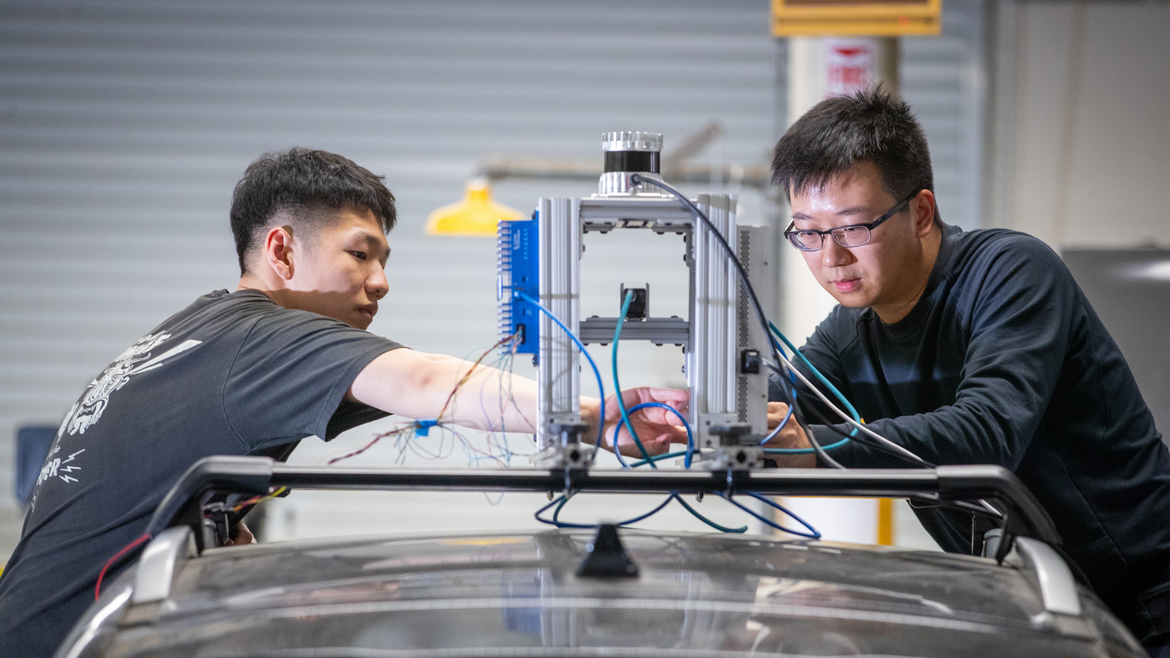Two researchers adjust a sensor-equipped apparatus mounted on top of a vehicle, working with exposed wiring and instrumentation in a laboratory setting.