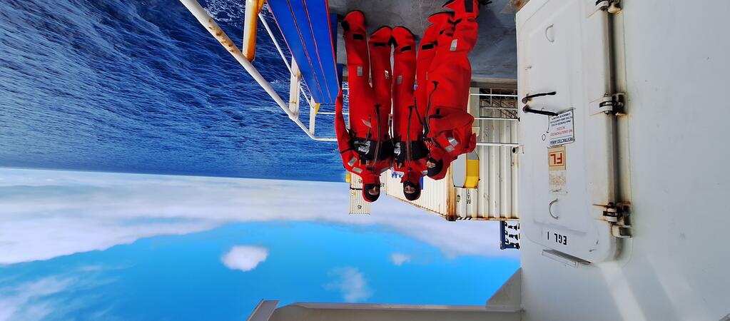 Three researchers wearing bright red immersion suits stand on the deck of a ship, with the open ocean and partly cloudy blue sky in the background. The group is positioned near a white metal door and railing, with containers and equipment visible on the deck.