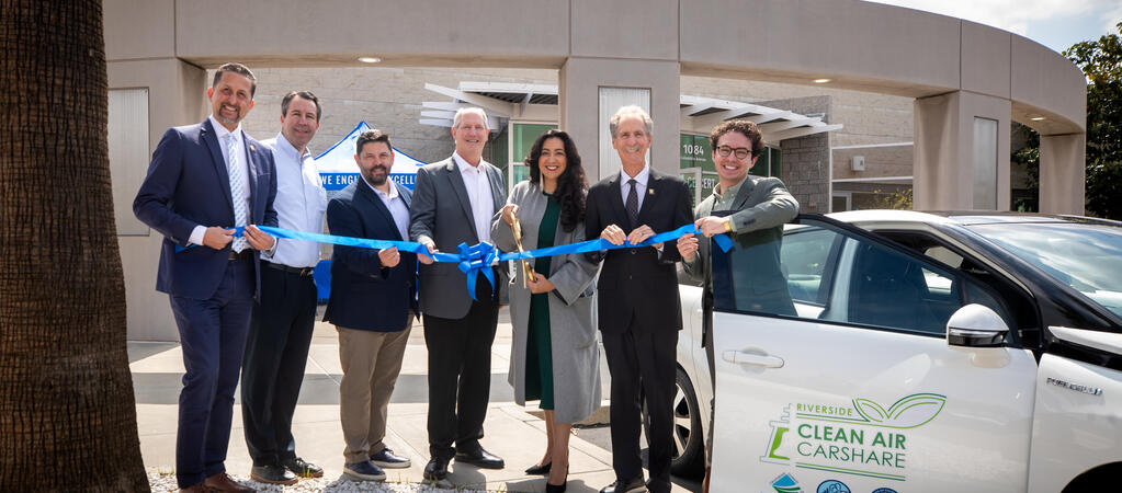 Eight individuals, including city officials, CE-CERT representatives, and project partners, stand in front of two hydrogen-powered carshare vehicles as they cut a large blue ceremonial ribbon to launch the Riverside Clean Air Carshare program at CE-CERT.