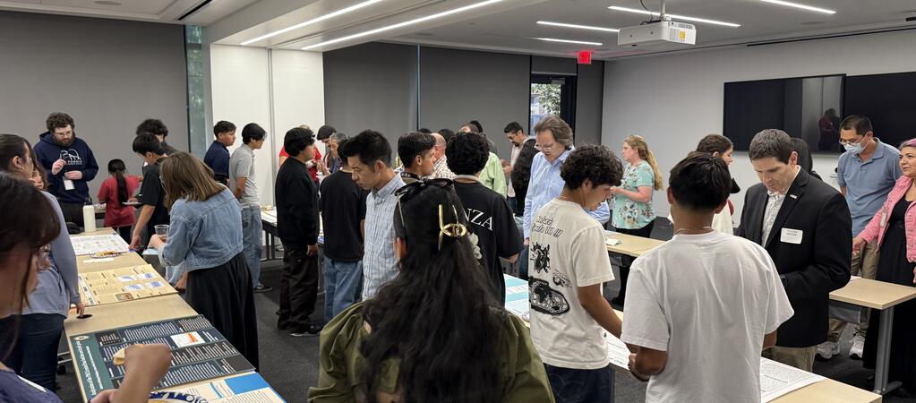 A classroom filled with students and adults viewing air quality research posters displayed on long tables. Attendees are reading, discussing, and pointing at various projects. The room is brightly lit with digital screens and projectors at the front. Don Collins is visible on the right side of the image, reviewing a student’s project.