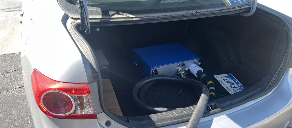 Rear view of a silver Toyota Corolla parked in a lot with a black emissions sampling hose extending from the trunk, indicating mobile emissions testing in progress.