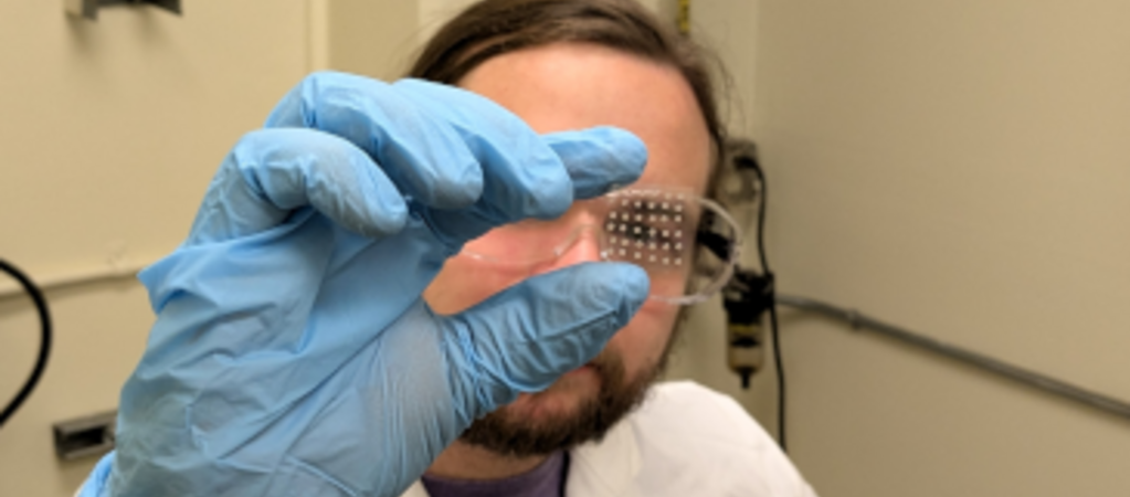 A researcher wearing blue gloves and a white lab coat holds up a transparent chip with small metallic dots, demonstrating ice nucleation research.