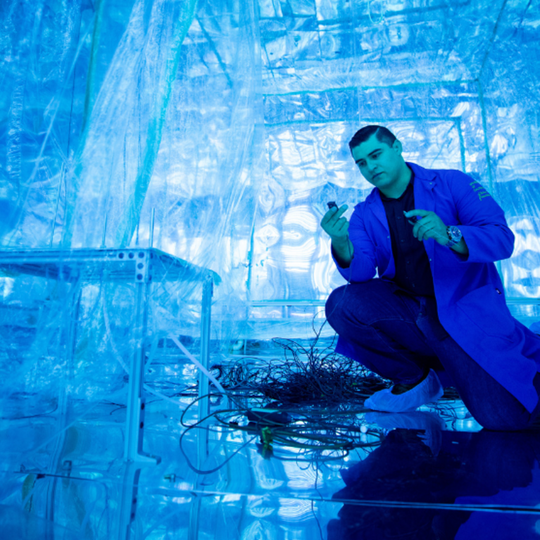 Researcher wearing a blue lab coat kneels inside the blue-lit Advanced Plant Laboratory (APL), inspecting sensors and wiring within a controlled-environment research chamber.