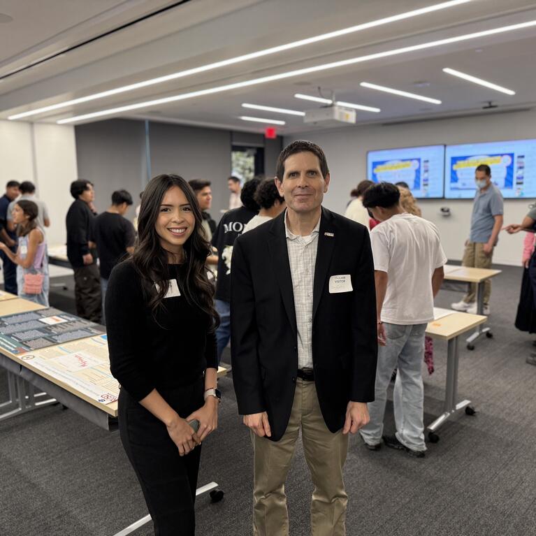 Kiara Salgado, a young woman with long dark hair wearing a black outfit, stands next to Don Collins, who is wearing a black blazer and khaki pants. They are both smiling at the camera in a classroom filled with students and poster presentations displayed on tables in the background.