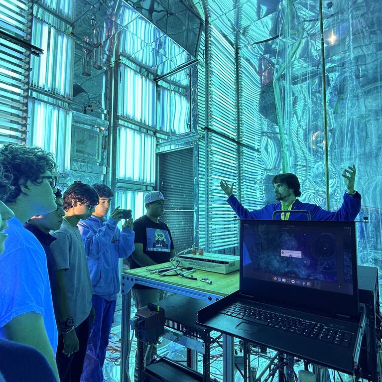 A group of five high school students on a STEPCon (Science and Technology Education Partnership Conference) tour are gathered in the world's largest indoor atmospheric processes chamber at the UC Riverside CE-CERT Atmospheric Processes Laboratory. The students are intently focused on a researcher in a blue jacket, who is enthusiastically explaining a concept with his arms raised, standing near a laptop. The massive chamber is dramatically lit by arrays of intense cyan/blue UV lights