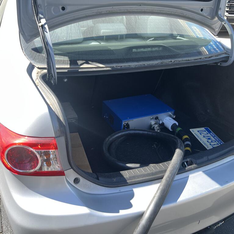 pen trunk of a silver Toyota Corolla showing a blue portable emissions analyzer connected to a black hose, used for measuring ammonia emissions during on-road testing.