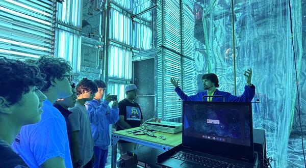 A group of five high school students on a STEPCon (Science and Technology Education Partnership Conference) tour are gathered in the world's largest indoor atmospheric processes chamber at the UC Riverside CE-CERT Atmospheric Processes Laboratory. The students are intently focused on a researcher in a blue jacket, who is enthusiastically explaining a concept with his arms raised, standing near a laptop. The massive chamber is dramatically lit by arrays of intense cyan/blue UV lights