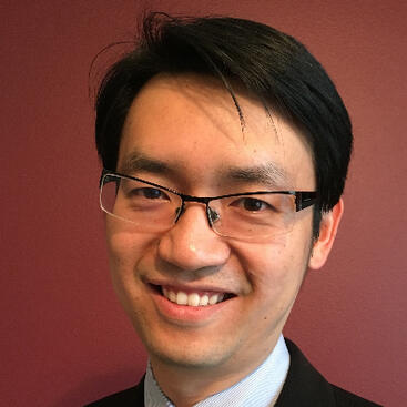 Portrait of Dr. Kanok Booriboonsoomsin wearing glasses, a suit, and a tie, smiling in front of a maroon background.