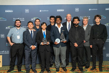 UCR EcoCAR team members stand together in formal and semi-formal attire, holding trophies in front of the official EcoCAR EV Challenge step-and-repeat backdrop featuring logos of GM, DOE, and MathWorks.