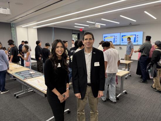 Kiara Salgado, a young woman with long dark hair wearing a black outfit, stands next to Don Collins, who is wearing a black blazer and khaki pants. They are both smiling at the camera in a classroom filled with students and poster presentations displayed on tables in the background.