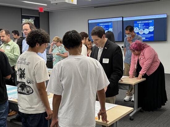 A group of students and adults are gathered in a classroom setting. Don Collins, wearing a black blazer and name tag, is standing at a table and reading a student’s project display. Other attendees are observing posters and engaging in conversations around the room.