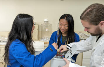 Two women and a man work together in a lab, with the women wearing blue lab coats and the man in a white lab coat, examining and discussing a piece of scientific equipment.