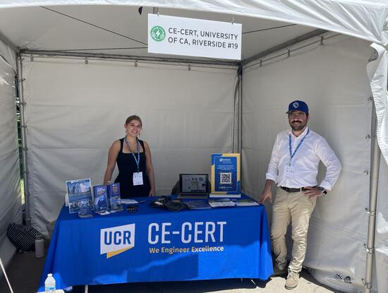 Two CE-CERT representatives stand behind an exhibit table displaying research materials and equipment inside a booth.