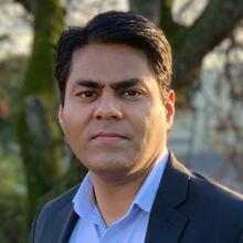 A man with short black hair, wearing a light blue collared shirt and dark blazer, looks at the camera while standing outdoors with a tree in the background.